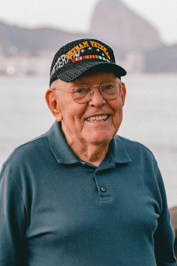 Elderly man wearing a Vietnam veteran cap, smiling near the ocean.